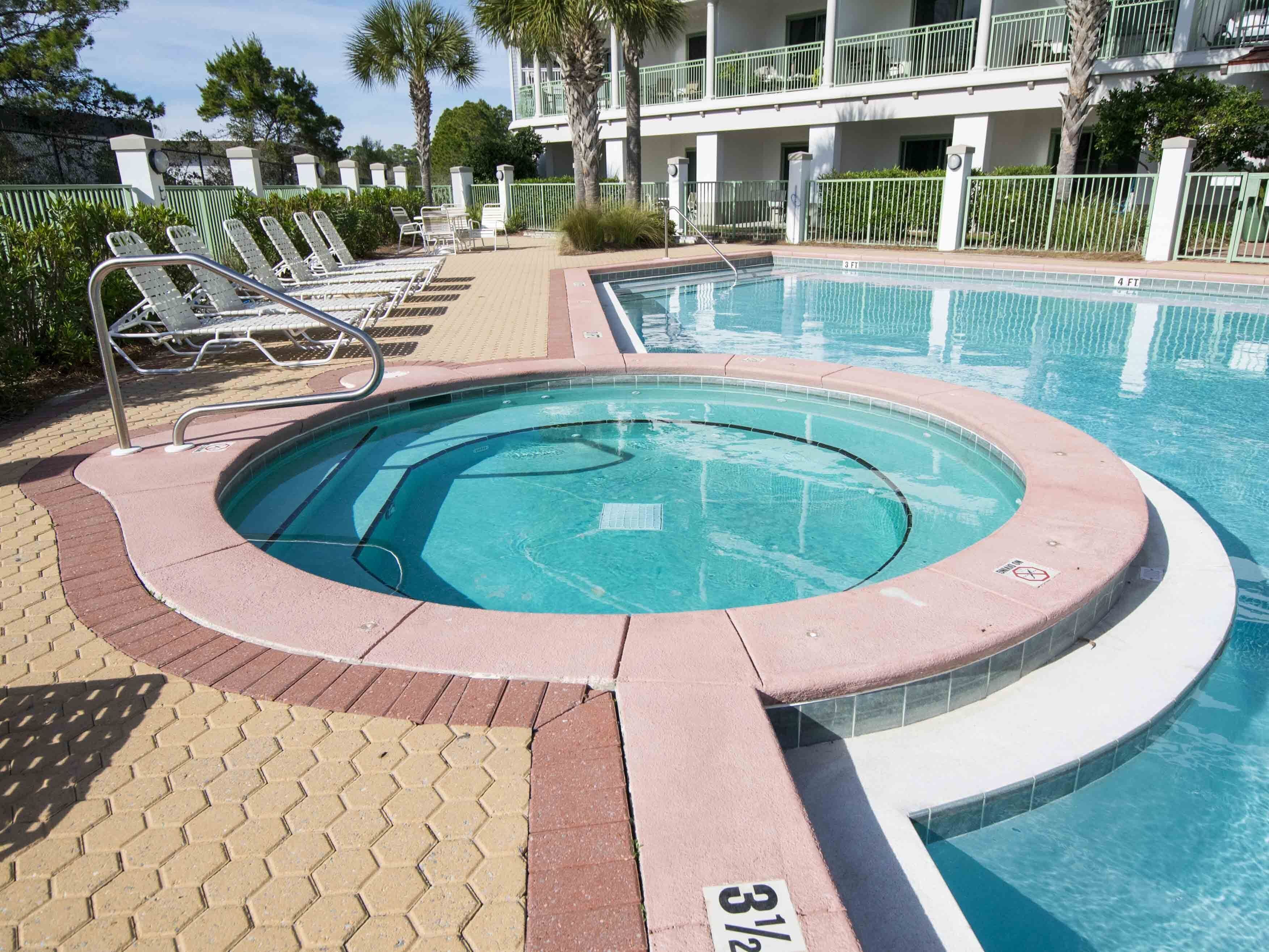 Una piscina con un'area idromassaggio circolare, lettini prendisole e un edificio con balconi sullo sfondo.
