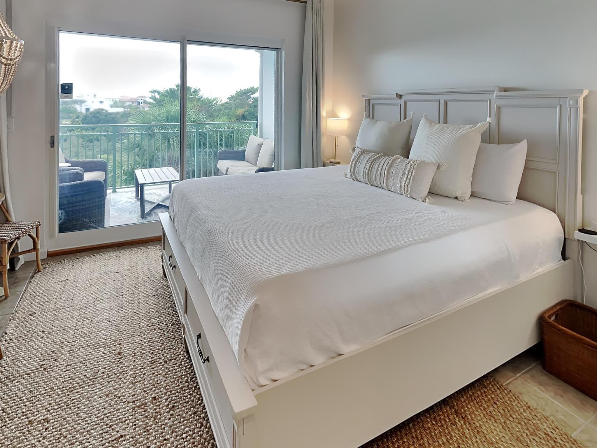 Ein großes Bett mit weißer Bettwäsche, ein Nachttisch, eine Lampe und Blick auf einen Balkon mit Gartenmöbeln.