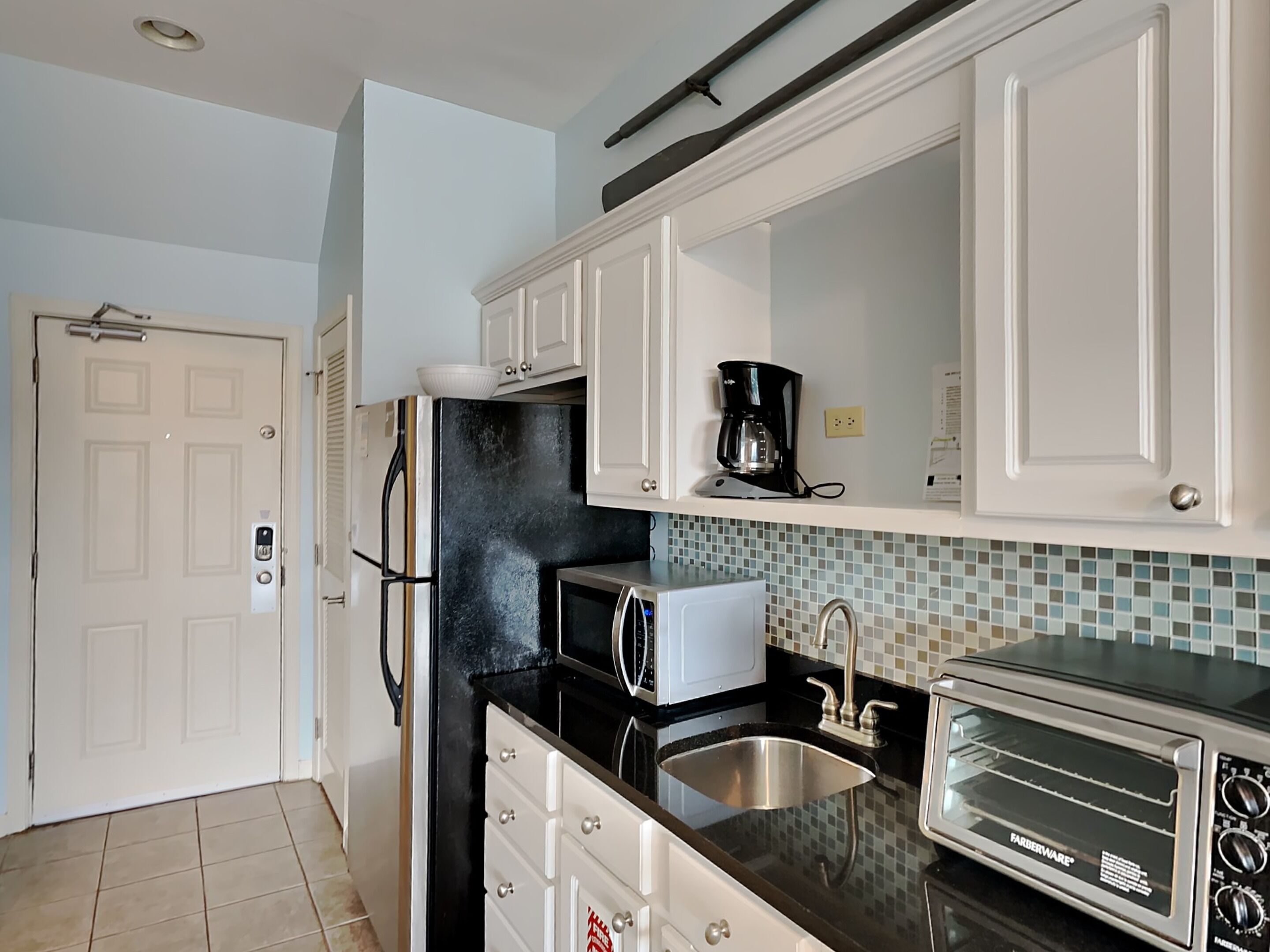 A kitchen with white cabinets, a black countertop, a stainless steel sink, a coffee maker, a microwave, and a toaster oven.