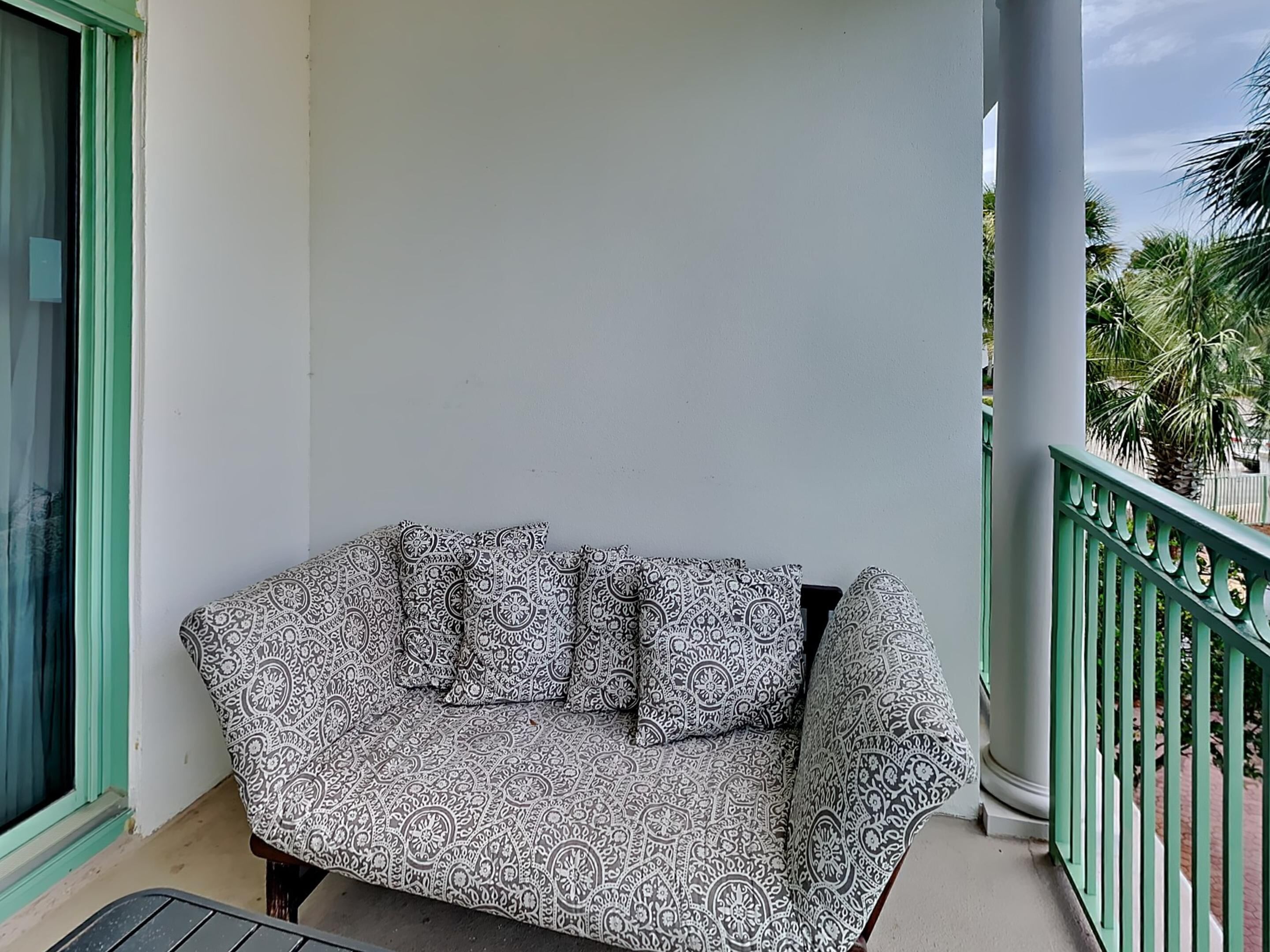En balkon med en stribet sofa og hynder, et grønt gelænder og udsigt til palmer.