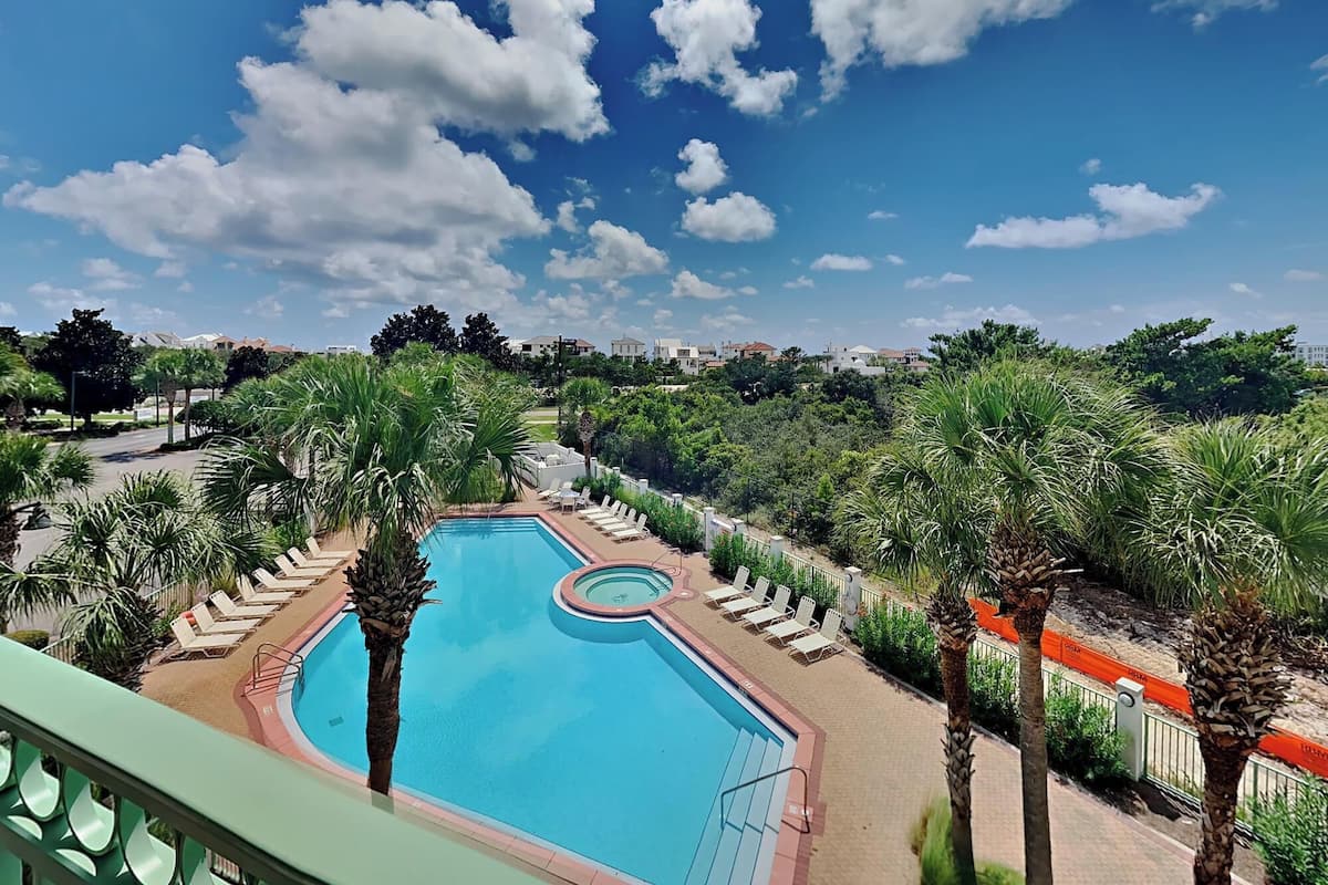 A swimming pool with lounge chairs and palm trees, surrounded by greenery and buildings in the background.