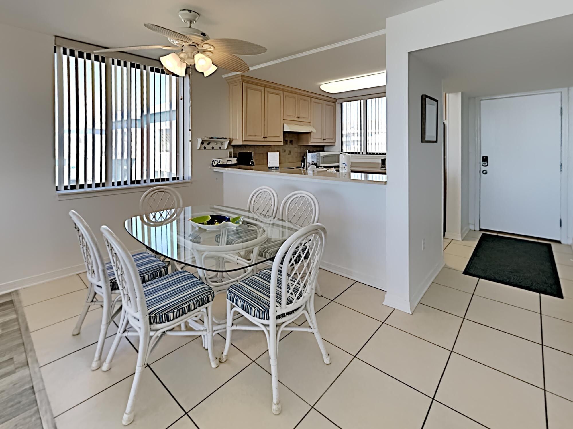 A modern kitchen with white cabinets, a dining area with a glass table and wicker chairs, and a ceiling fan.