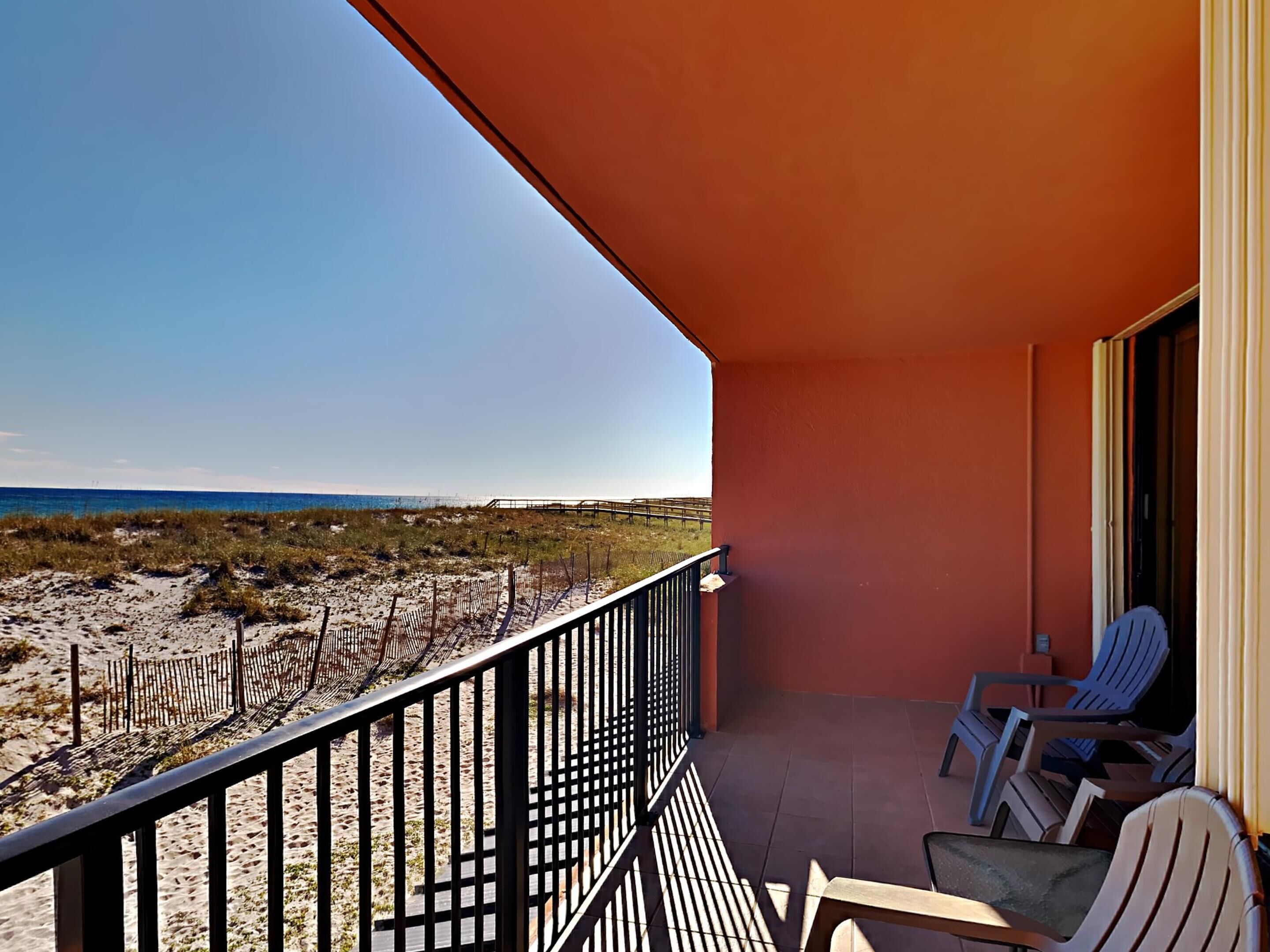 A balcony with a view of the beach, featuring a railing, a few chairs, and a door.
