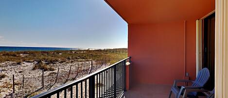 A balcony with a view of the beach, featuring a railing, a few chairs, and a door.