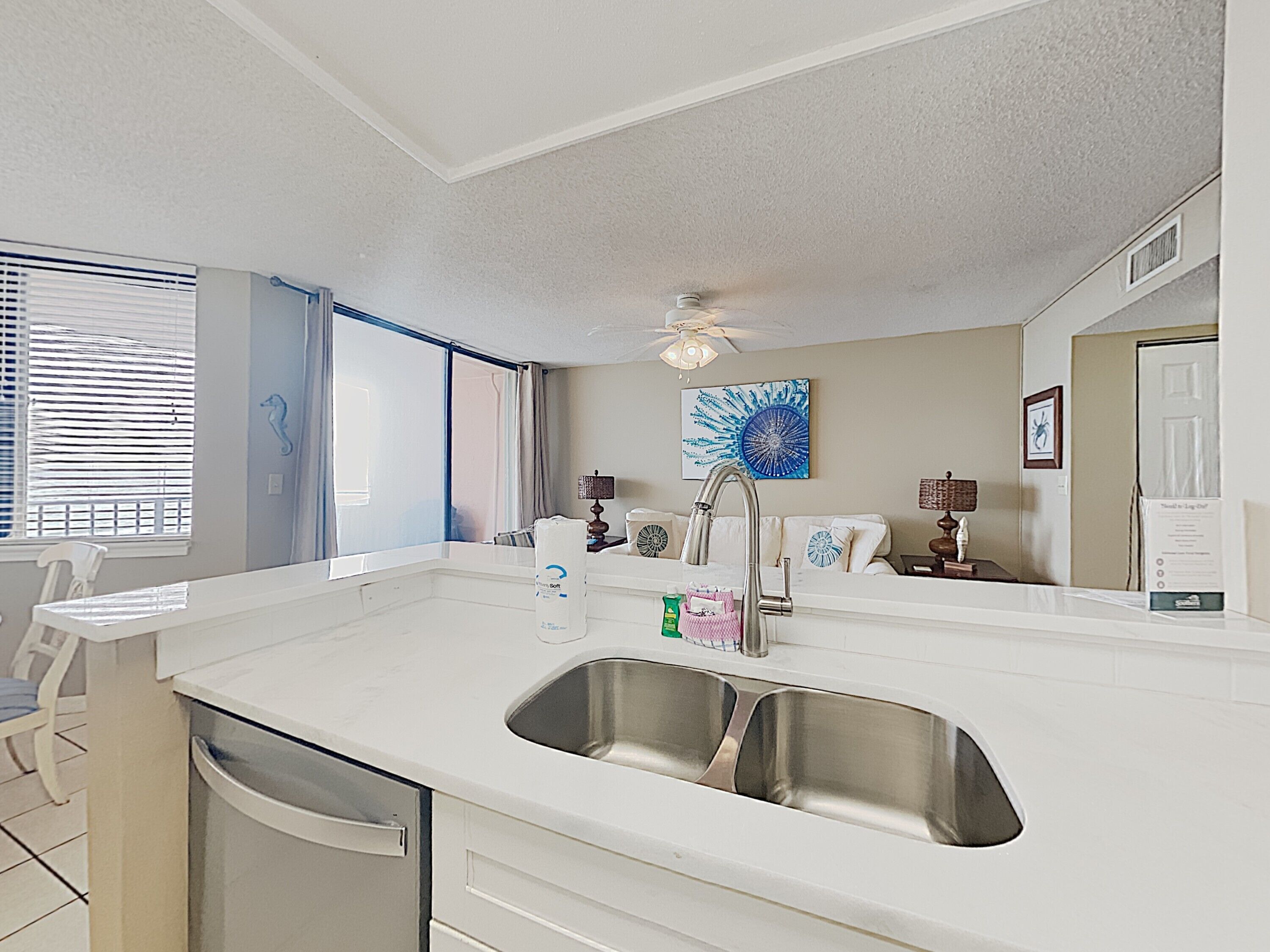 A modern kitchen with a white countertop, two stainless steel sinks, a ceiling fan, and a window with blinds.