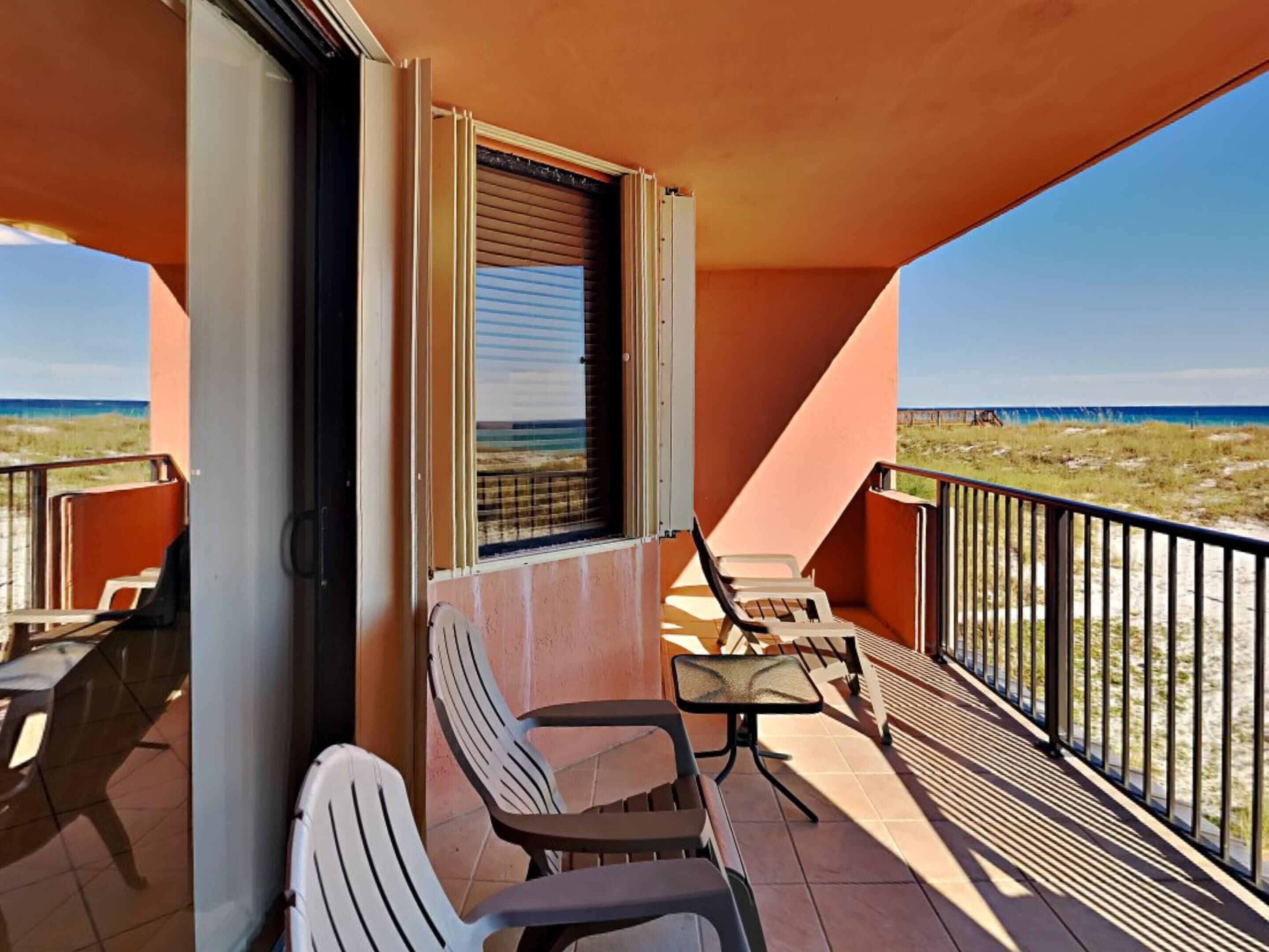 Ein Balkon mit zwei Stühlen und einem Tisch, der auf einen Strand mit blauem Himmel blickt.