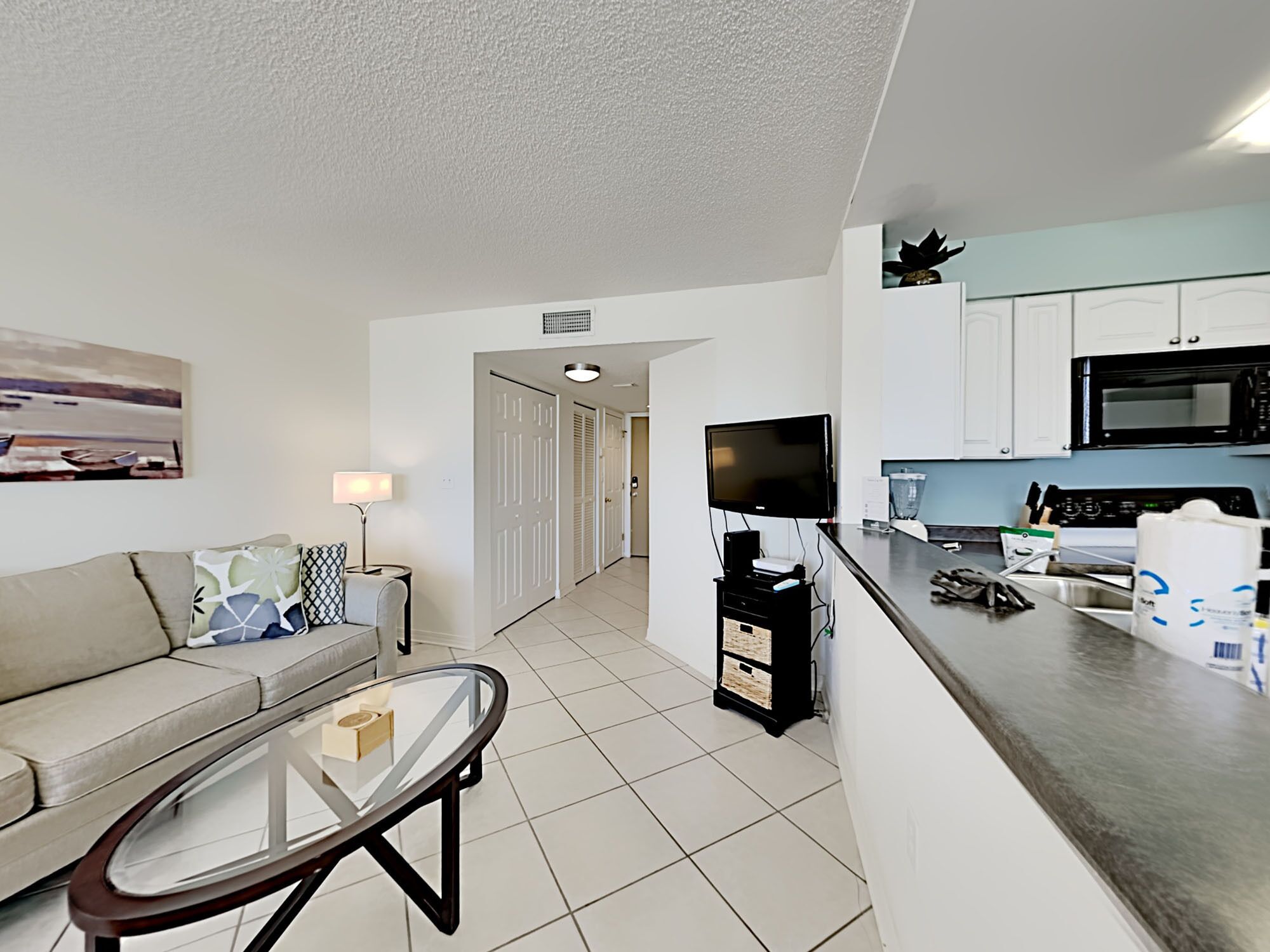 A modern living room with a sofa, a glass coffee table, a TV, and a kitchen area with a microwave and cabinets.