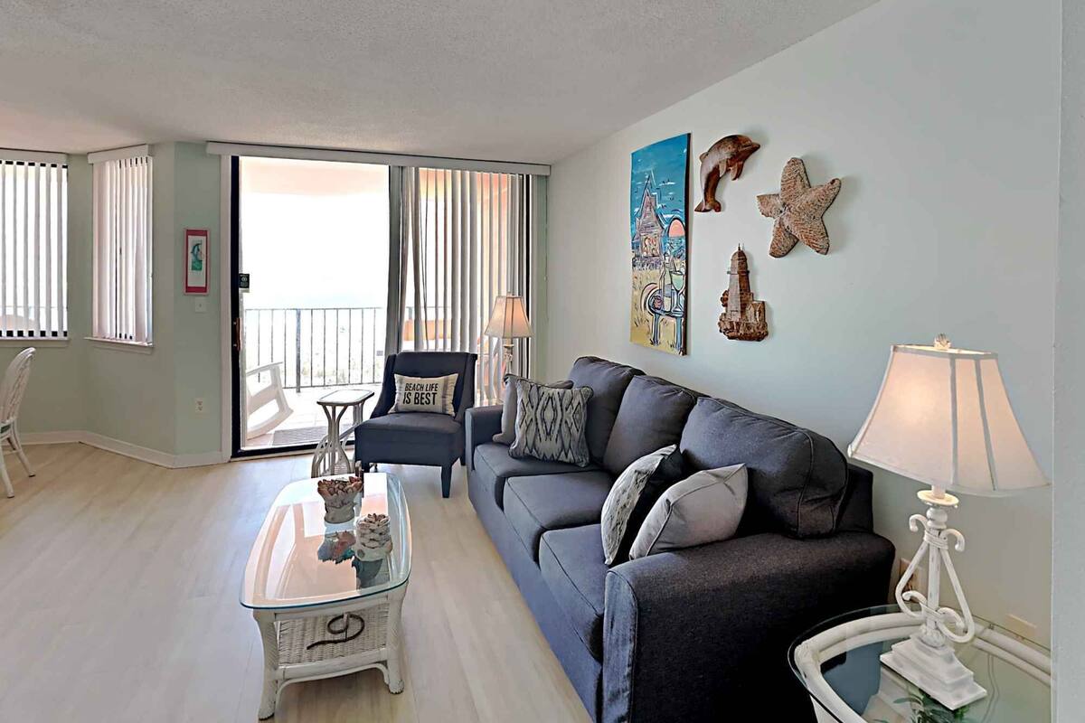 Un salon avec un canapé gris foncé, une table basse en verre, un fauteuil blanc et une petite table d’appoint avec une lampe. Deux tableaux ornent le mur et une fenêtre est dotée de stores blancs.