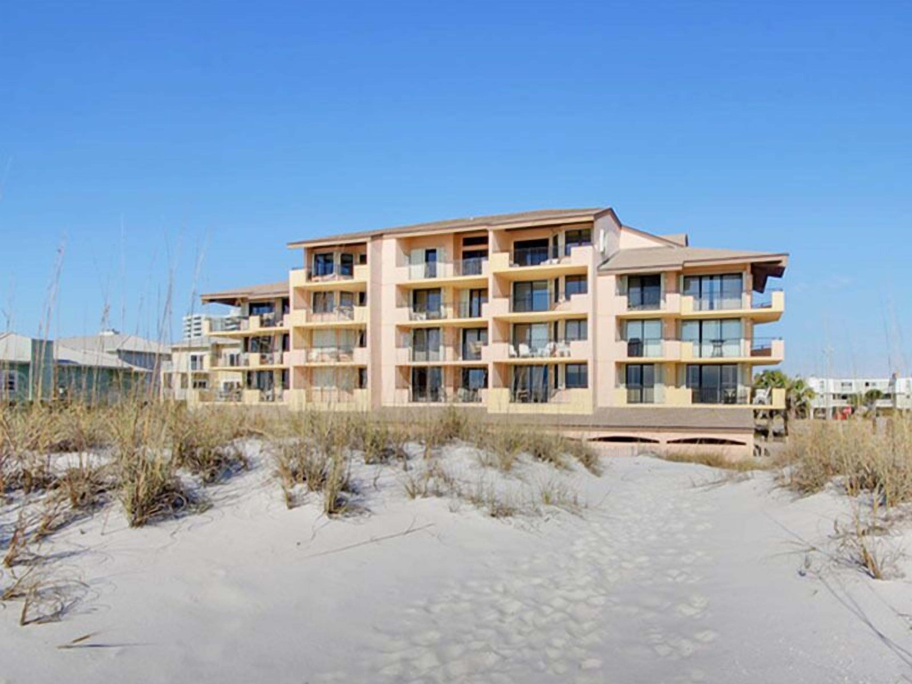 A multi-story hotel with balconies, located on a sandy beach with sparse vegetation.