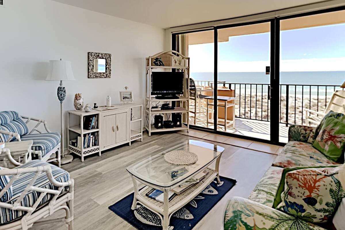 Un salon avec une table basse en verre, des meubles en osier, un meuble pour la télévision et une vue sur la plage grâce à de grandes portes coulissantes.