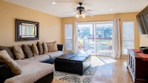 A living room with a sofa, ottoman, ceiling fan, and a view through glass doors to an outdoor patio.