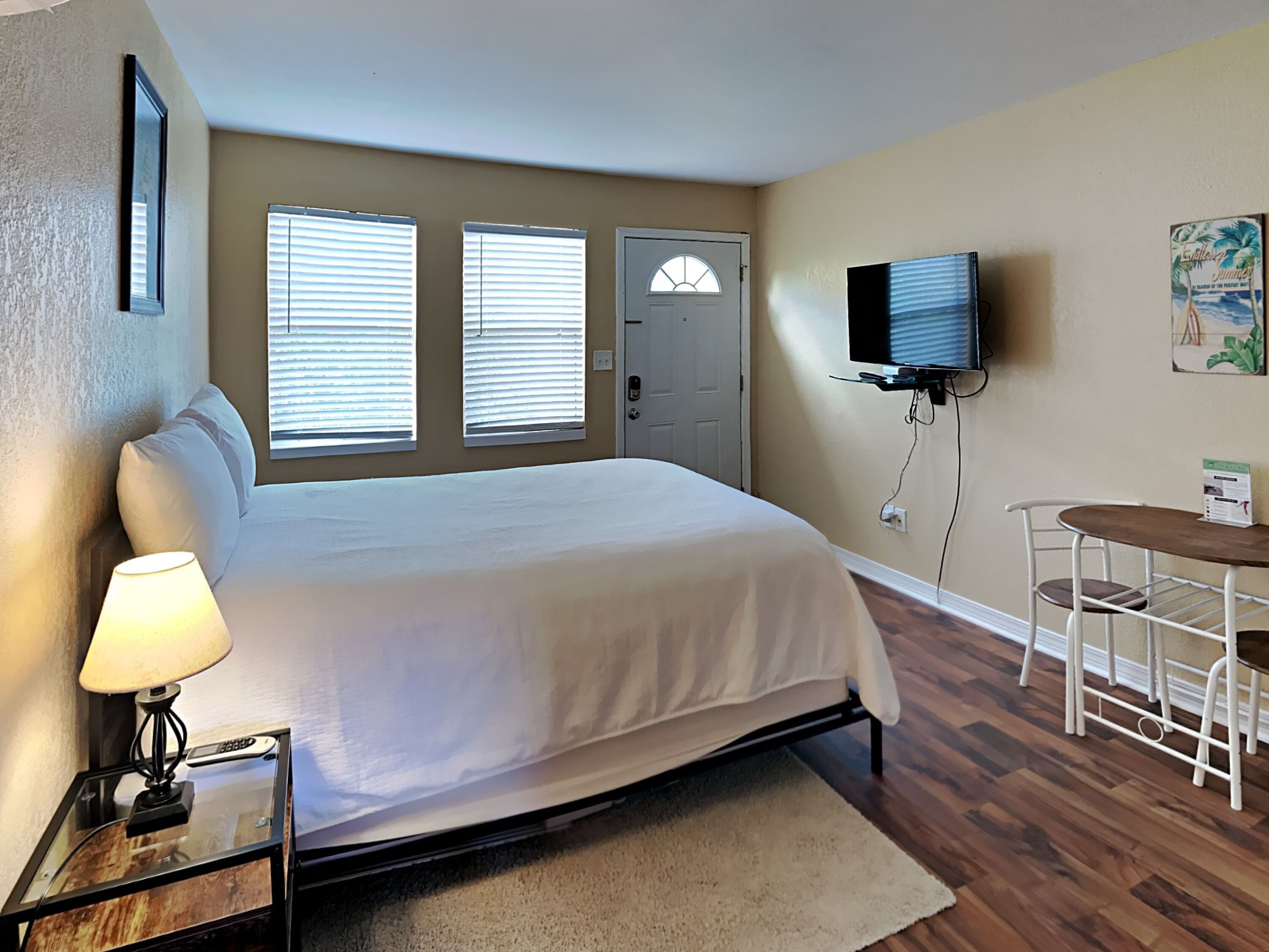 A bedroom with a bed, bedside table with a lamp, a TV mounted on the wall, a door, and a small table with a chair.