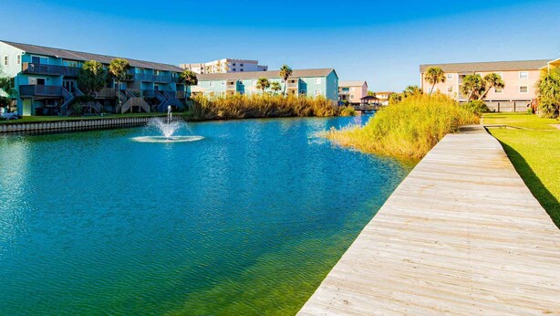 A waterfront property with a wooden boardwalk, a fountain, and buildings with balconies and palm trees.