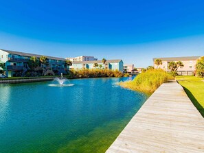 A waterfront property with a wooden boardwalk, a fountain, and buildings with balconies and palm trees. - Moonlight Beach Villa by Southern Vacation Rentals (Pensacola Beach)