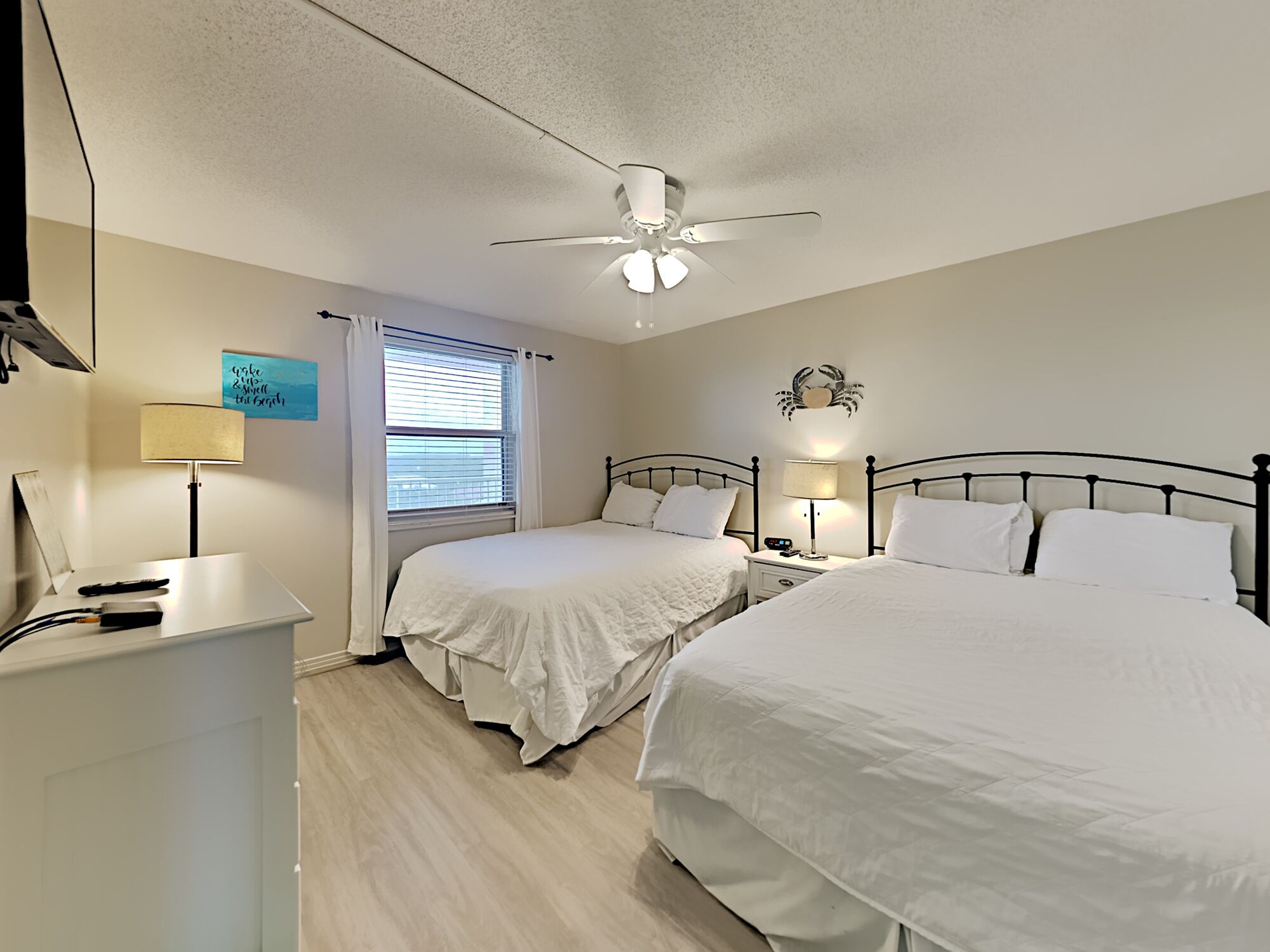 A bedroom with two beds, a ceiling fan, a bedside table with a lamp, and a window with blinds.