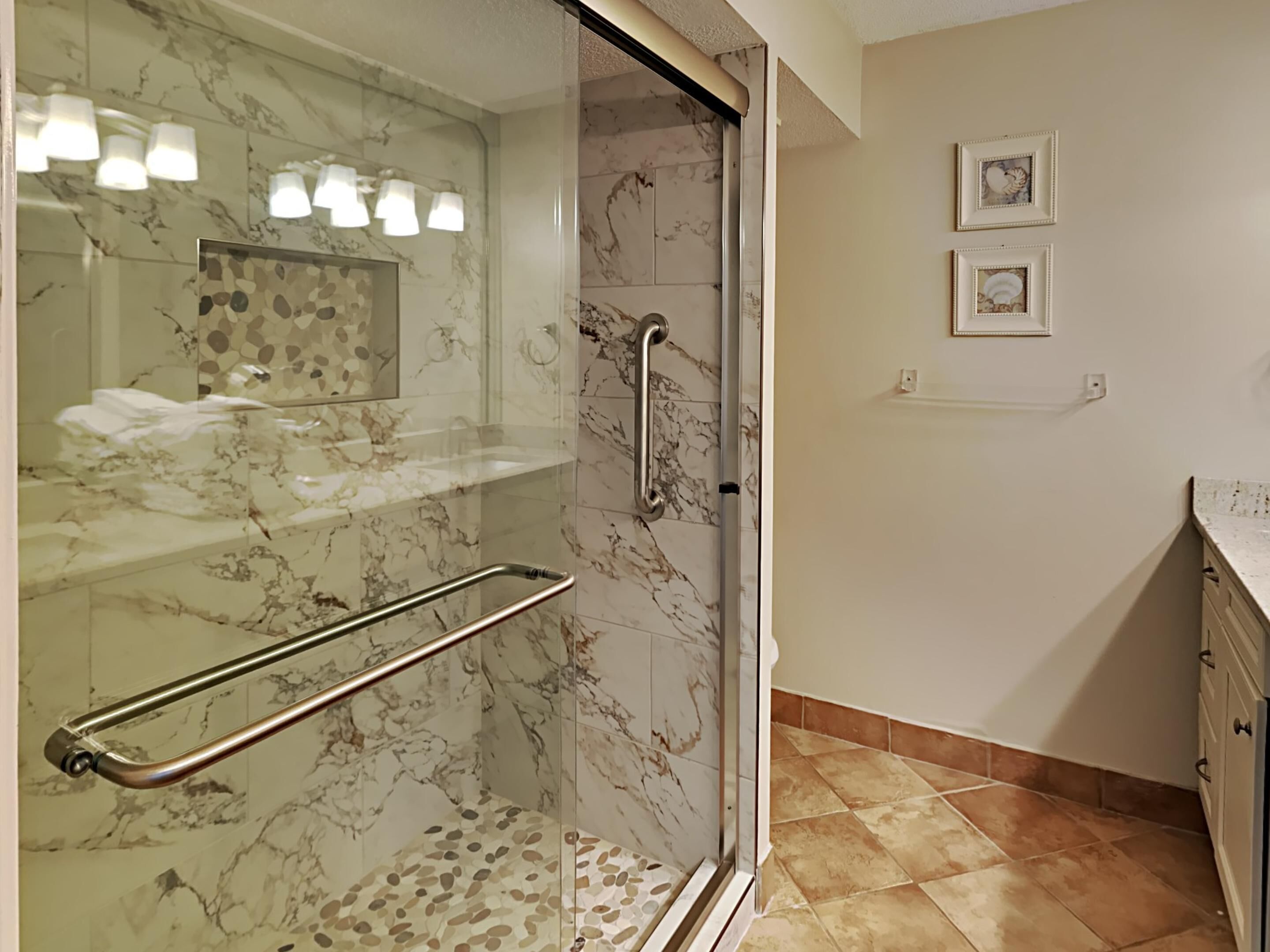 A modern bathroom with a glass shower enclosure, marble tiles, a handrail, and a small sink.