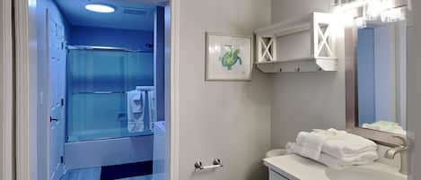 A bathroom with a glass shower enclosure, a white sink with a mirror, and a framed picture on the wall.