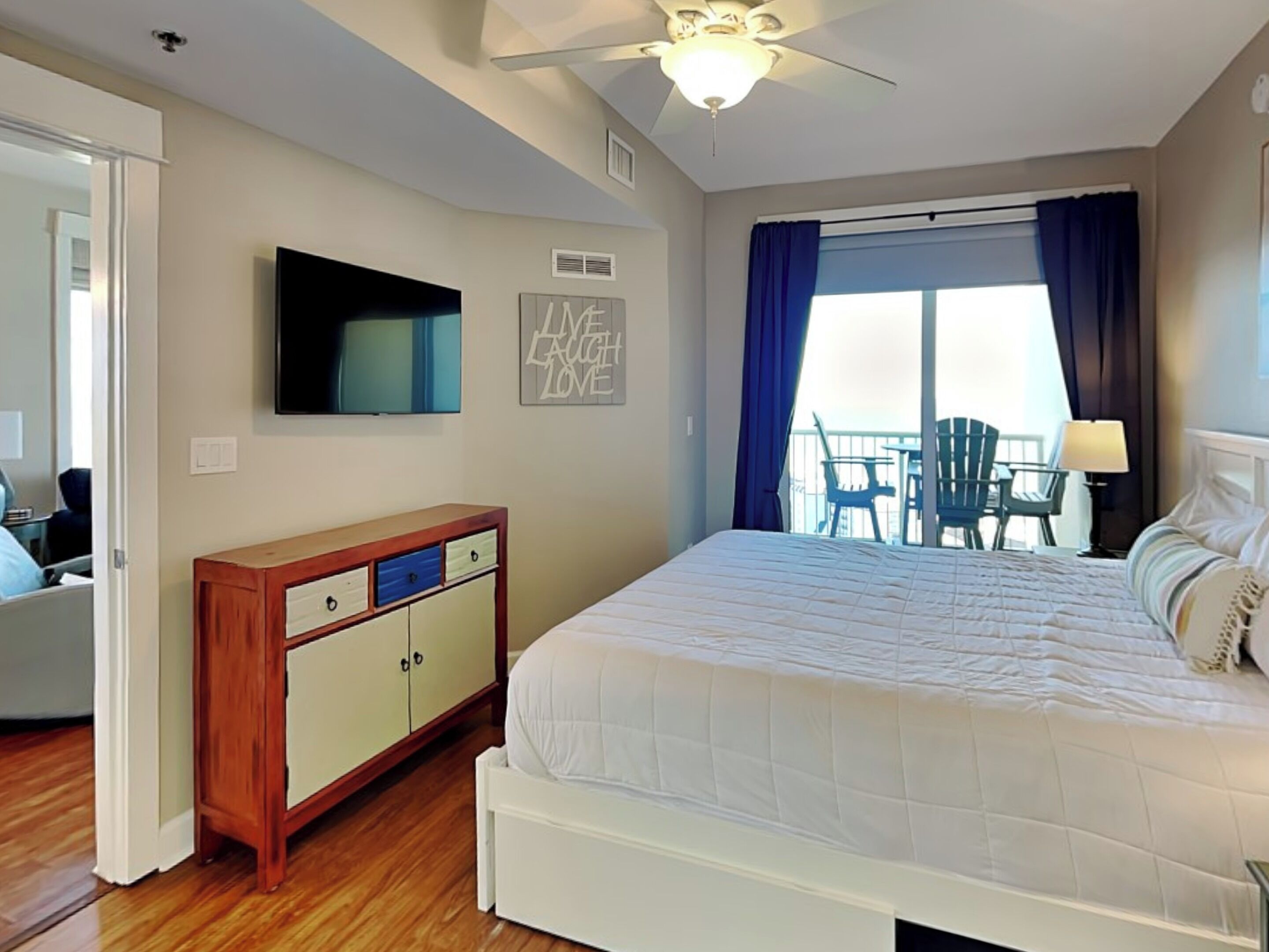 A hotel room with a bed, a TV, a wooden dresser, and a balcony with chairs.