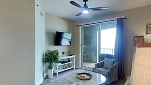 Una sala de estar con televisor de pantalla plana, ventilador de techo, puerta de vidrio que da a un balcón y una mesa de centro redonda.