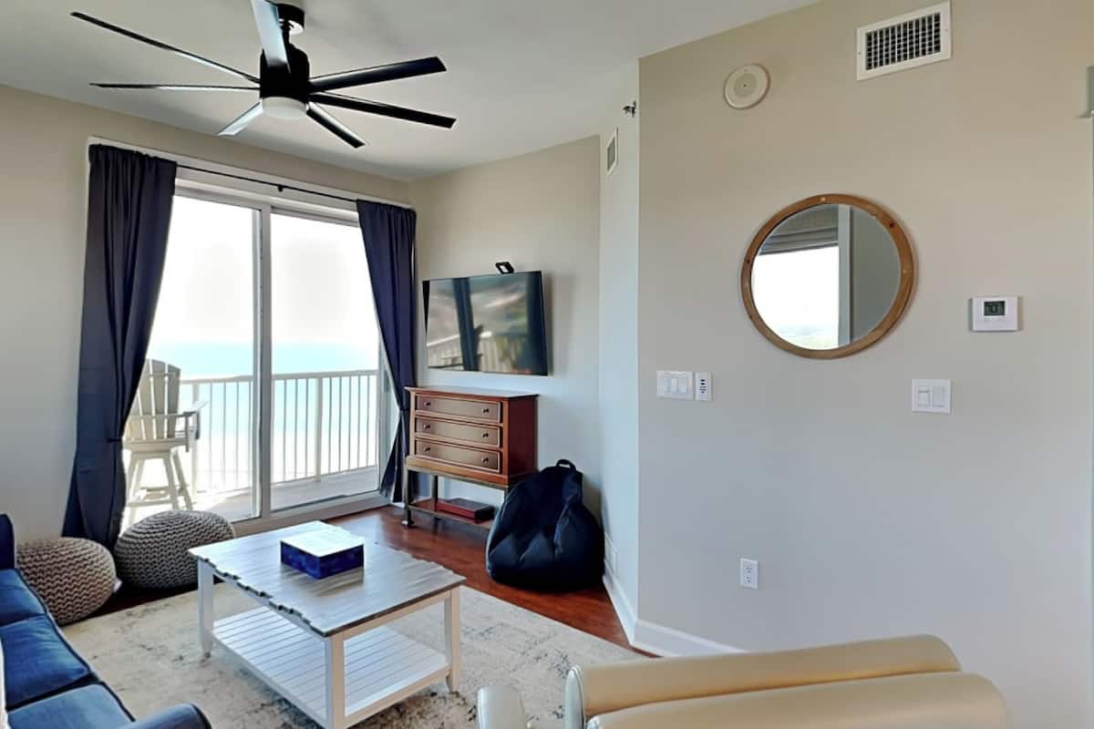 A living room with a sofa, a coffee table, a TV, and a balcony with a view.