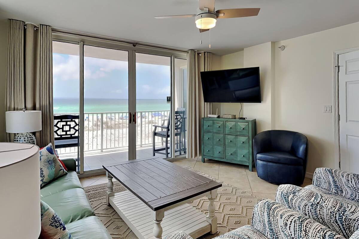 Ein Zimmer mit einer Couch, einem Couchtisch, einem Fernseher auf einem Schrank, einem Deckenventilator und Blick auf den Strand.