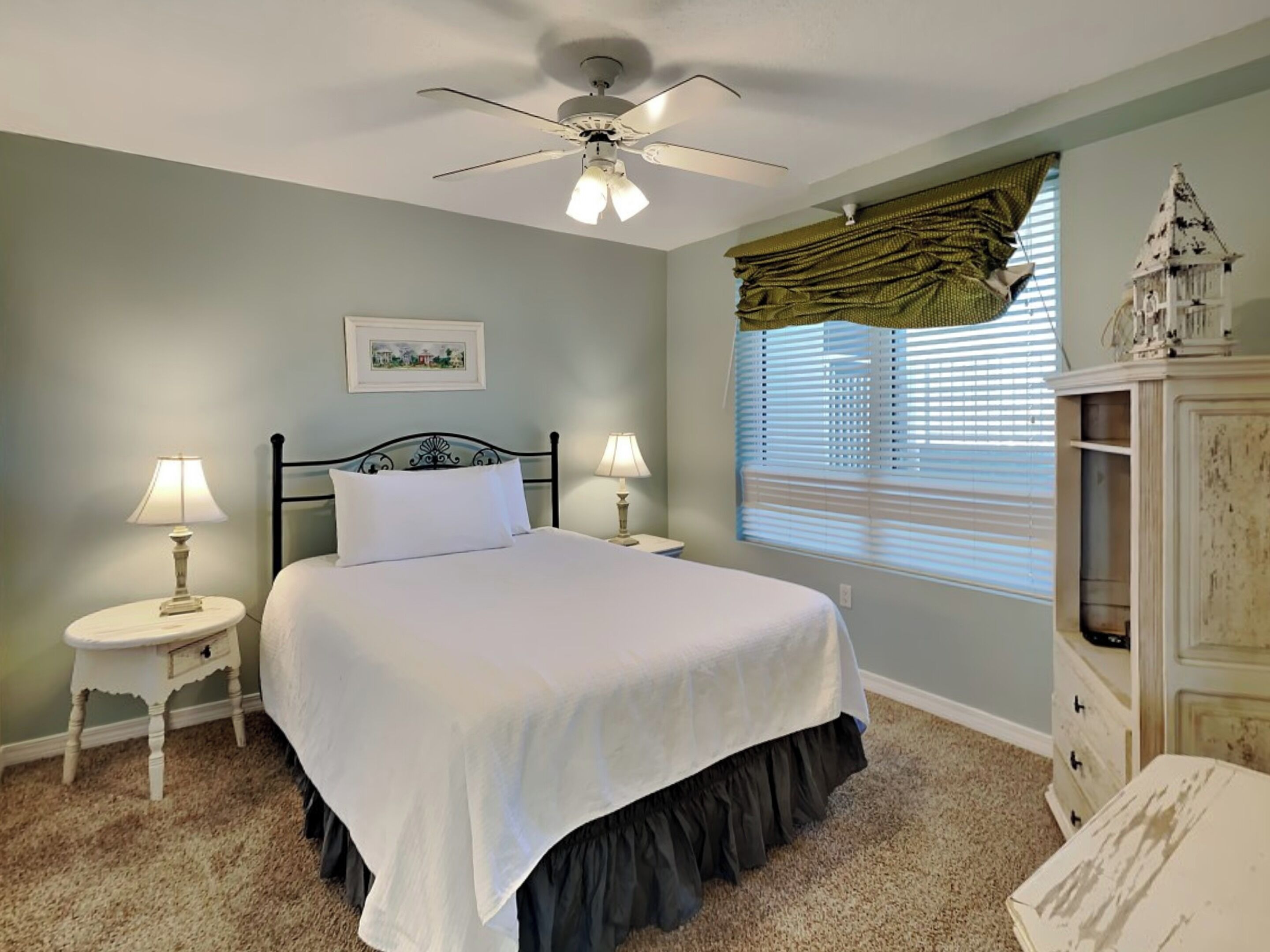 A bedroom with a bed, two bedside tables with lamps, a ceiling fan, a window with blinds, and a dresser.