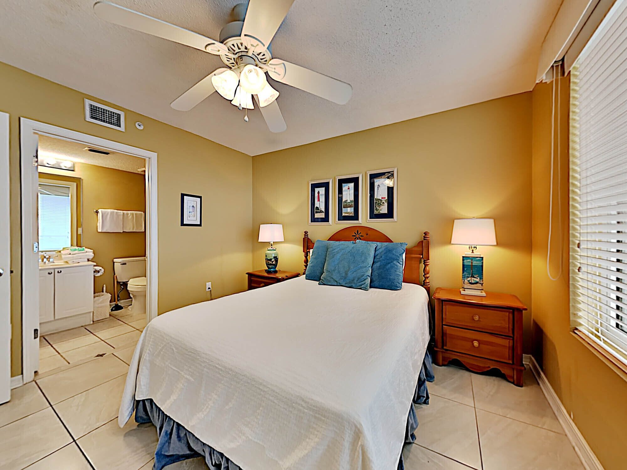 A hotel room with a bed, two bedside tables with lamps, a ceiling fan, a bathroom with a toilet and sink, and framed pictures on the wall.