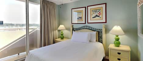 A hotel room with a bed, two bedside lamps, a ceiling fan, a large window with curtains, and two framed pictures on the wall.