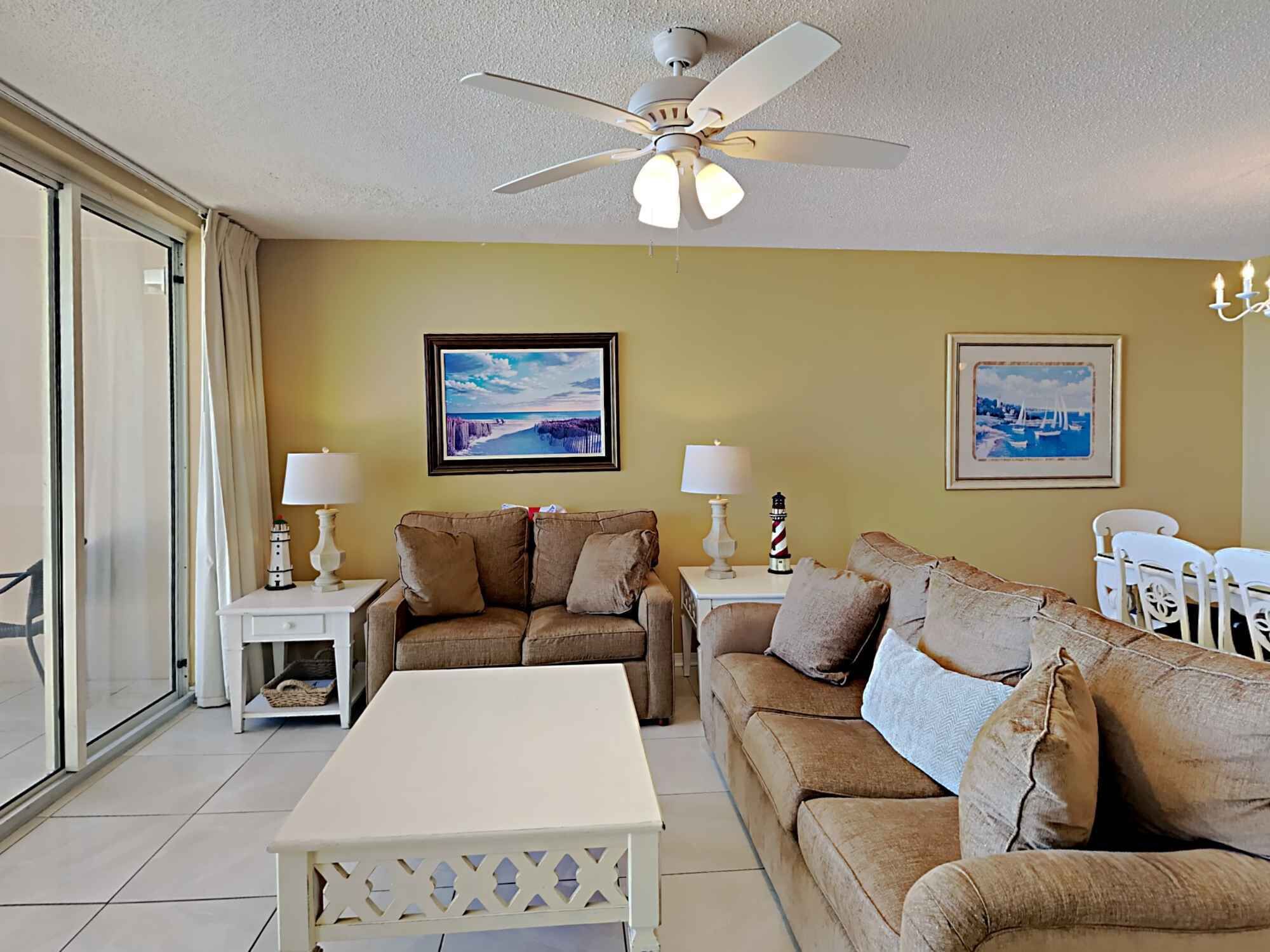A living room with a beige sofa, a white coffee table, and a ceiling fan. There are two framed pictures on the wall and a lamp on each side of the sofa.