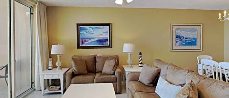 A living room with a beige sofa, a white coffee table, and a ceiling fan. There are two framed pictures on the wall and a lamp on each side of the sofa.