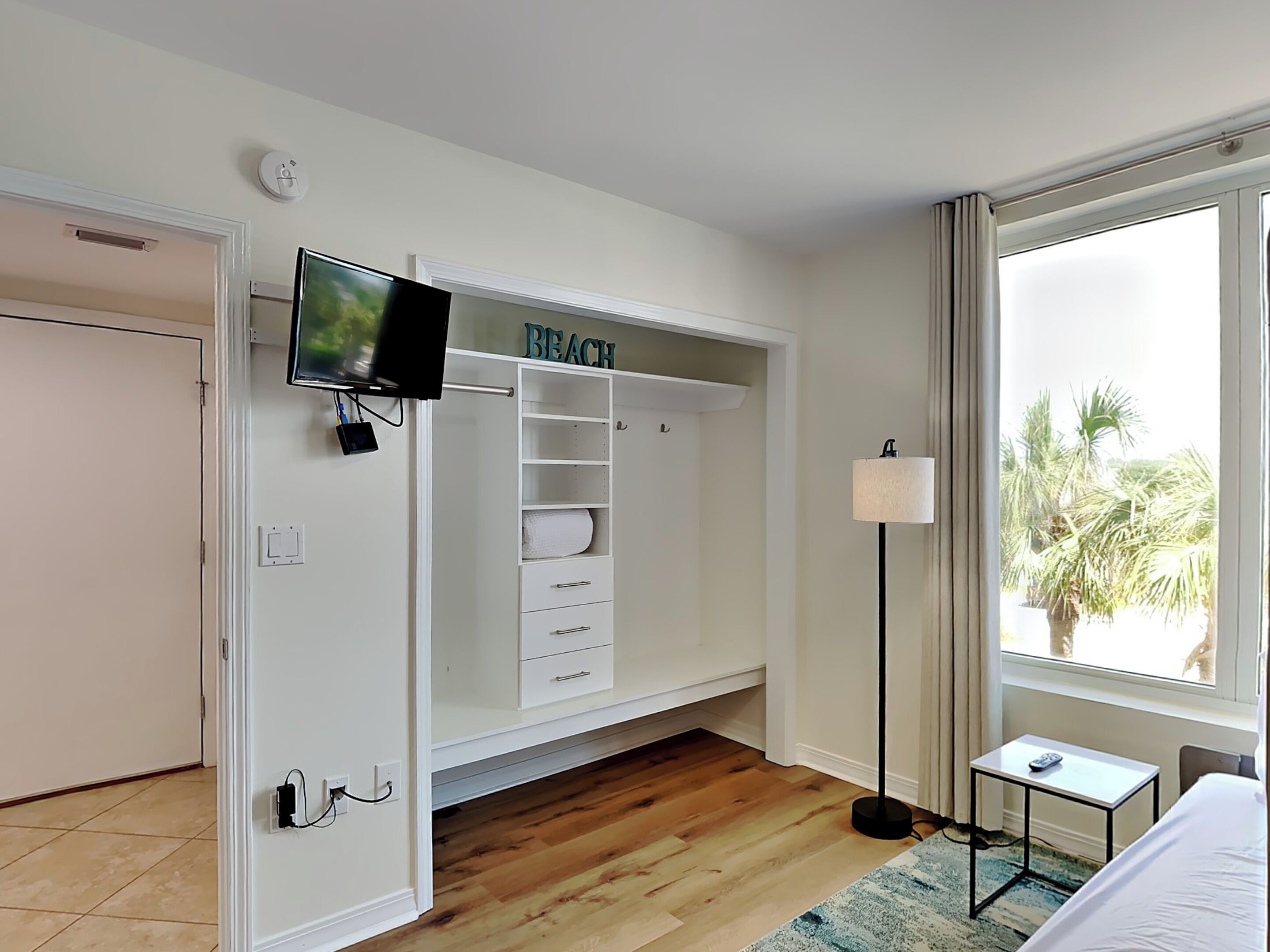 Hotelkamer met een aan de muur bevestigde televisie, een kast met een 'BEACH' bord, een nachtkastje met een lamp en een groot raam met uitzicht op palmbomen.