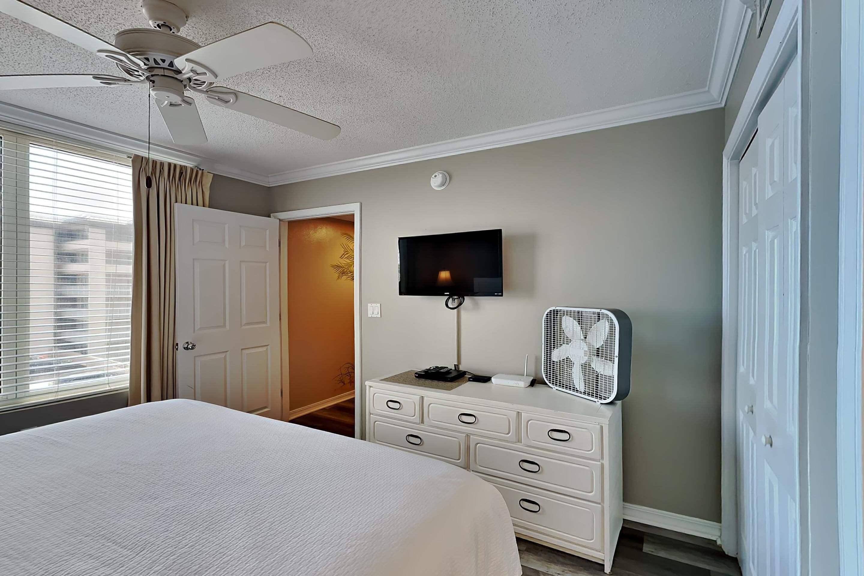 A bedroom with a bed, a ceiling fan, a TV, a dresser, and a fan on the dresser.