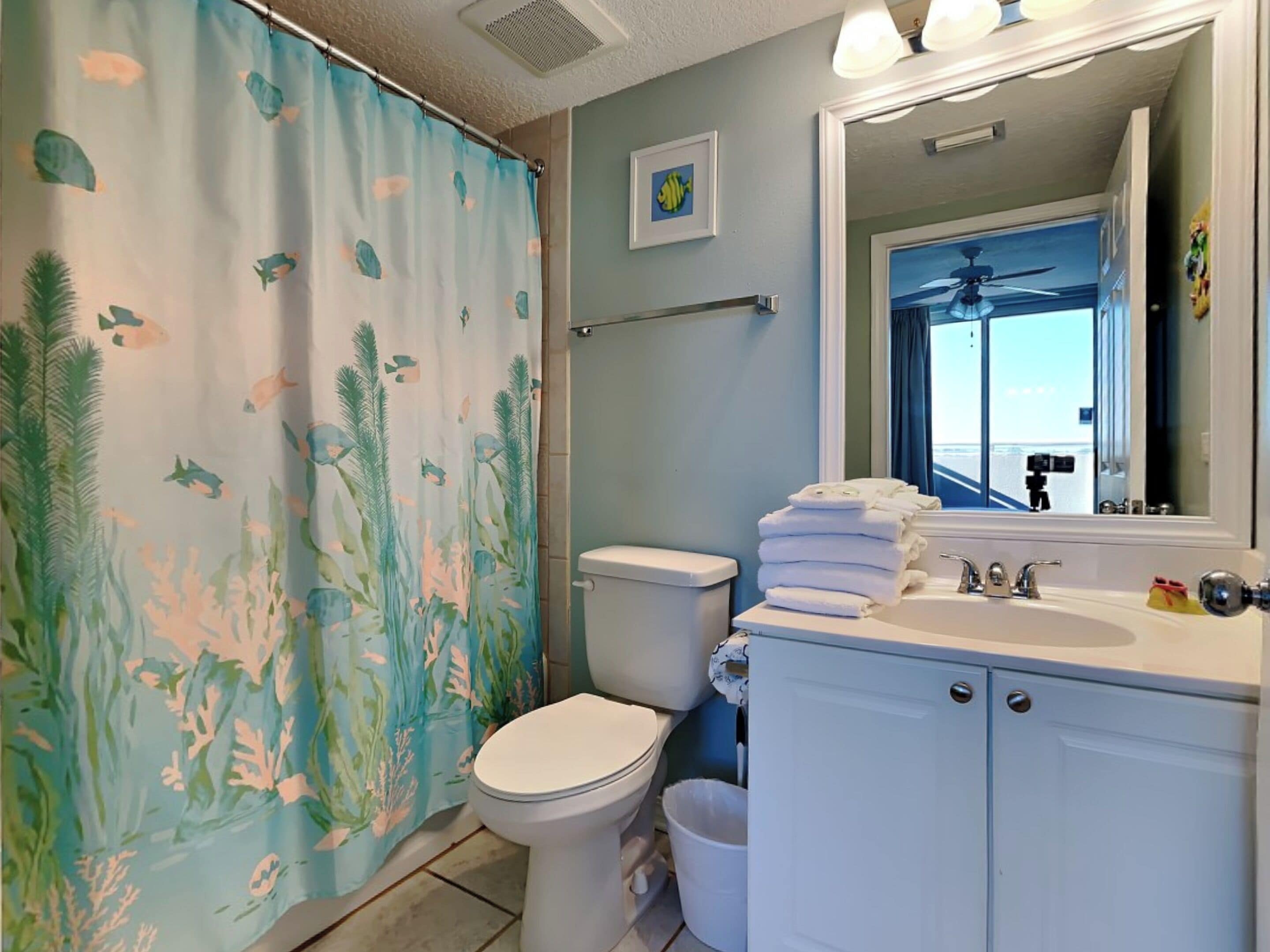 A bathroom with a shower curtain featuring a marine life pattern, a toilet, a sink with stacked towels, and a mirror reflecting a ceiling fan.