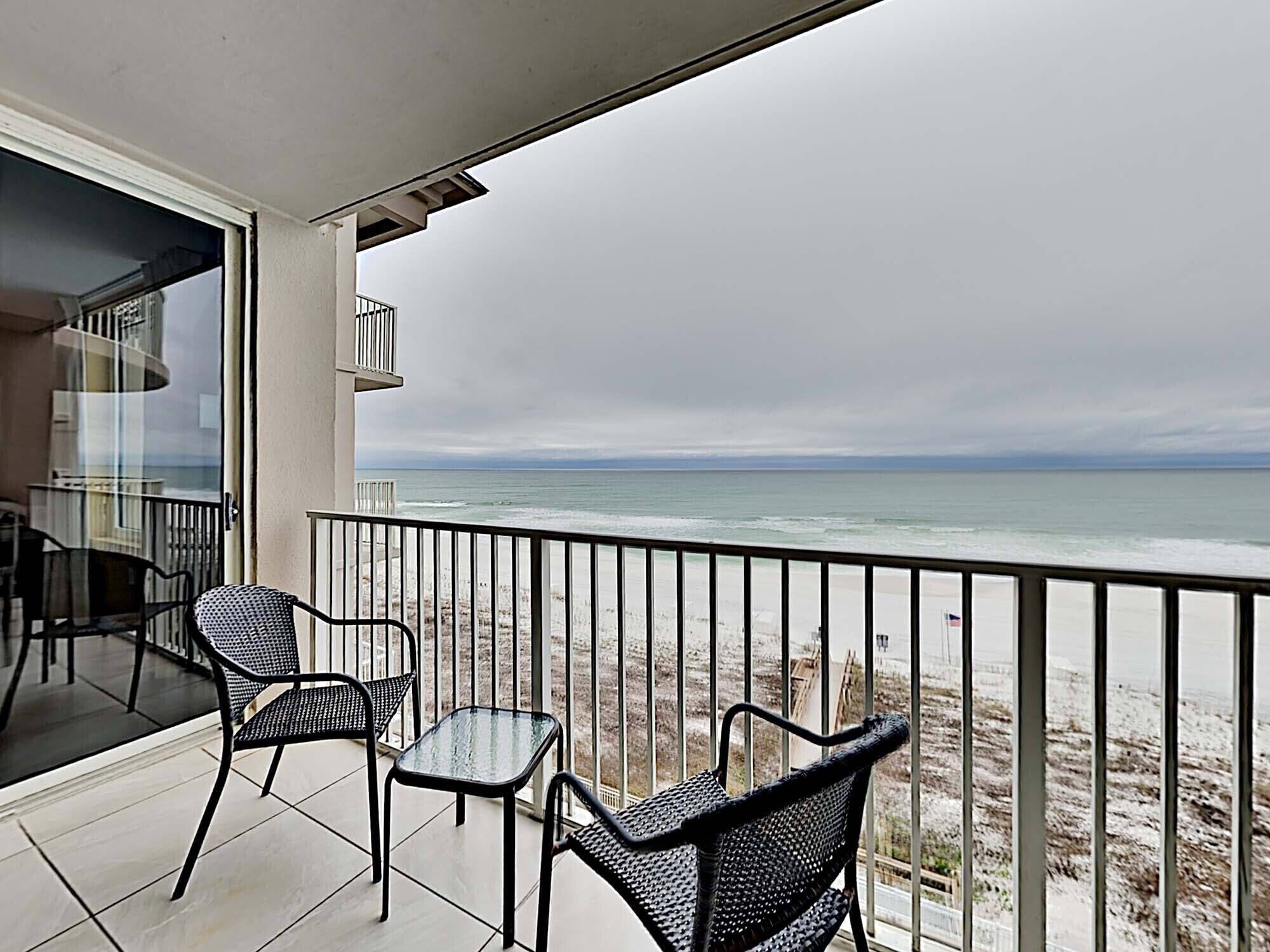 A balcony with two wicker chairs and a small table, overlooking a beach with a railing and a cloudy sky.