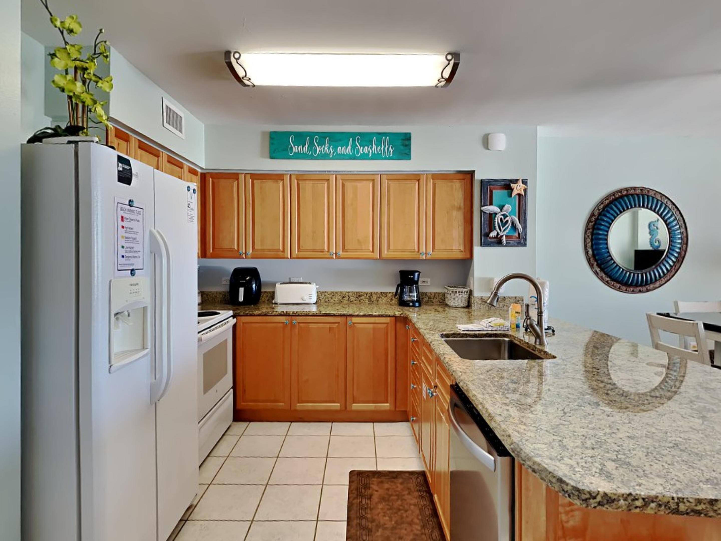 A kitchen with wooden cabinets, a granite countertop, a refrigerator, a coffee maker, and a sign that reads "Sand, Seas, and Shells".