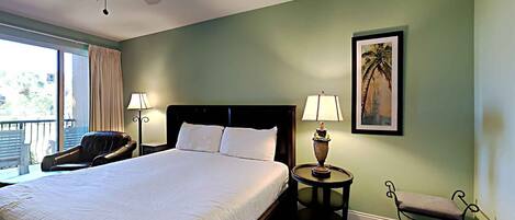 A hotel room with a large bed, a ceiling fan, a chair, a small table with a lamp, and a balcony with a chair.