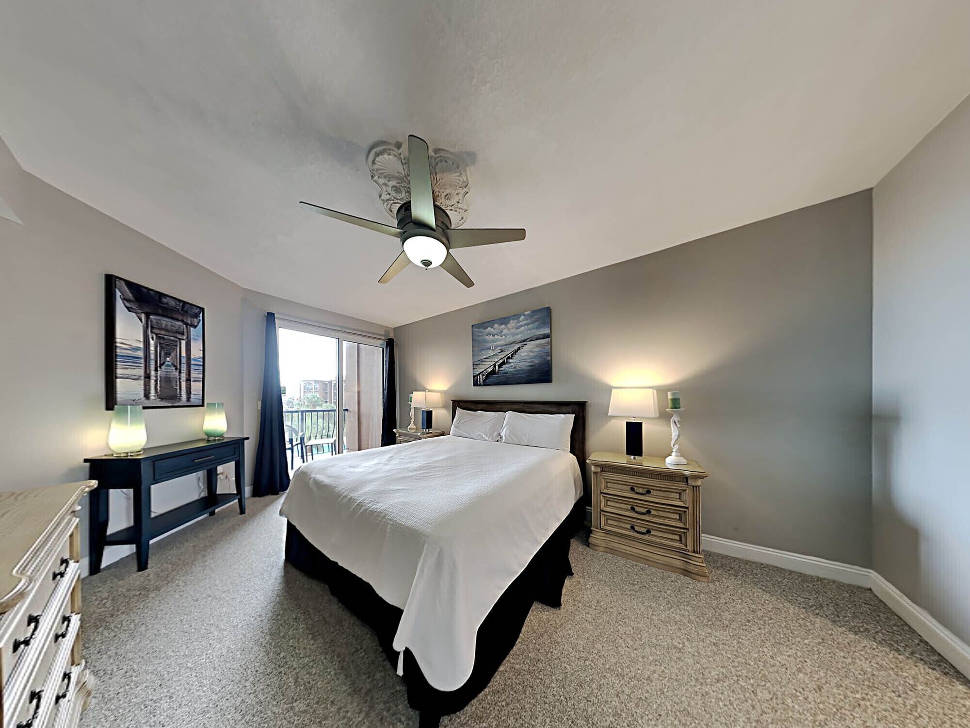 A hotel room with a large bed, a ceiling fan, a nightstand with a lamp, a dresser, and a painting on the wall.