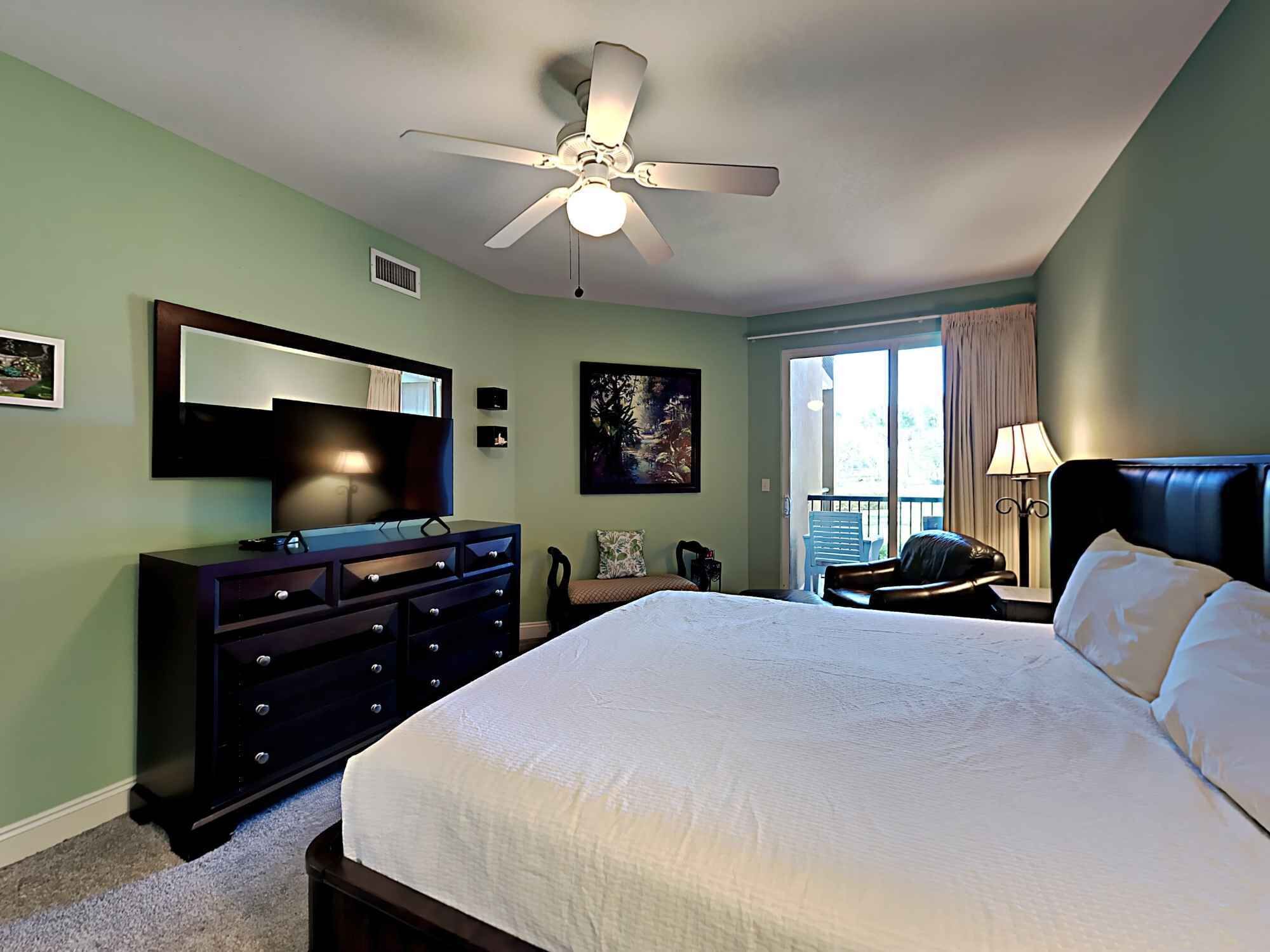A bedroom with a large bed, a ceiling fan, a dresser, a TV, and a window with curtains.