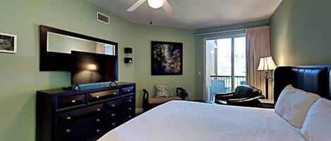 A bedroom with a large bed, a ceiling fan, a dresser, a TV, and a window with curtains.