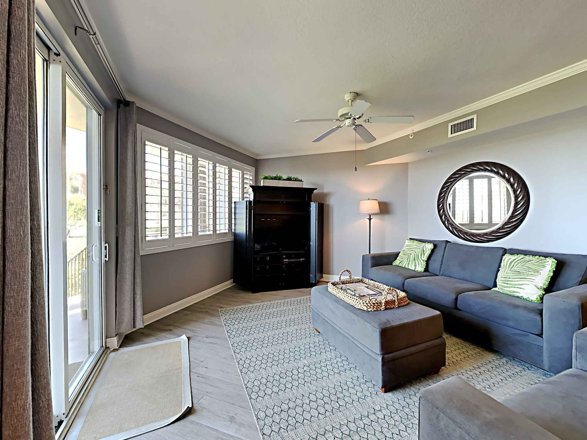 A living room with a grey sofa, a ceiling fan, a fireplace, and a large window.
