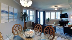 A modern living room with a glass dining table, wooden chairs, a chandelier, a ceiling fan, and a TV. - Mediterranea by Southern Vacation Rentals (Miramar Beach)