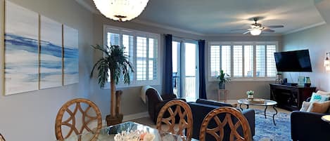 A modern living room with a glass dining table, wooden chairs, a chandelier, a ceiling fan, and a TV.
