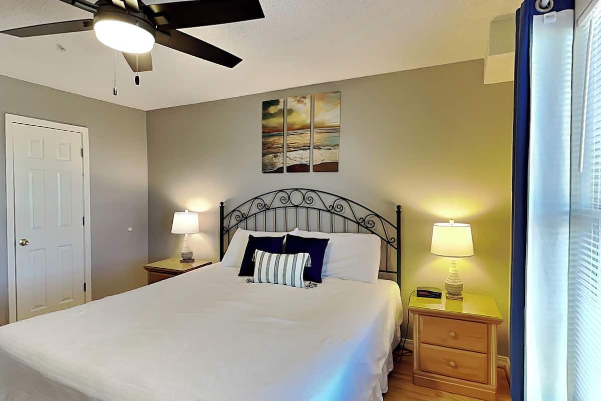 A neatly made bed with a headboard, two bedside tables with lamps, a ceiling fan, and a painting on the wall.