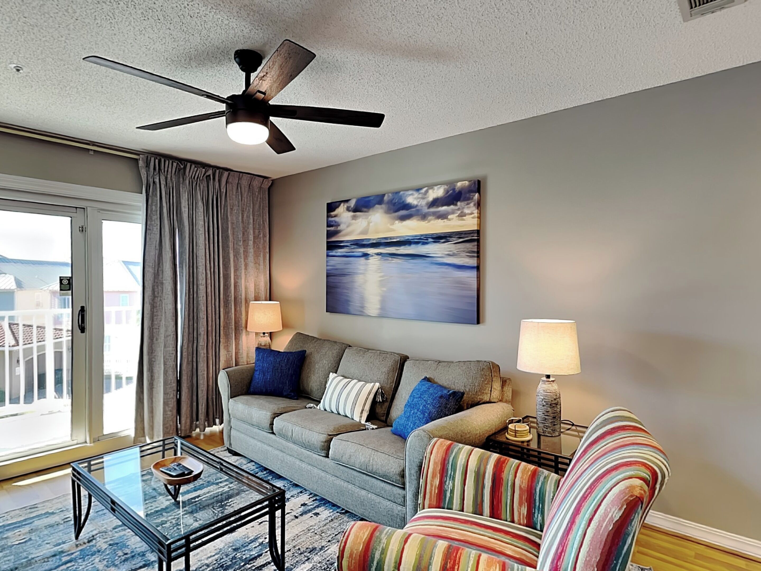 A living room with a gray sofa, striped armchair, glass coffee table, ceiling fan, and a painting of a beach scene.