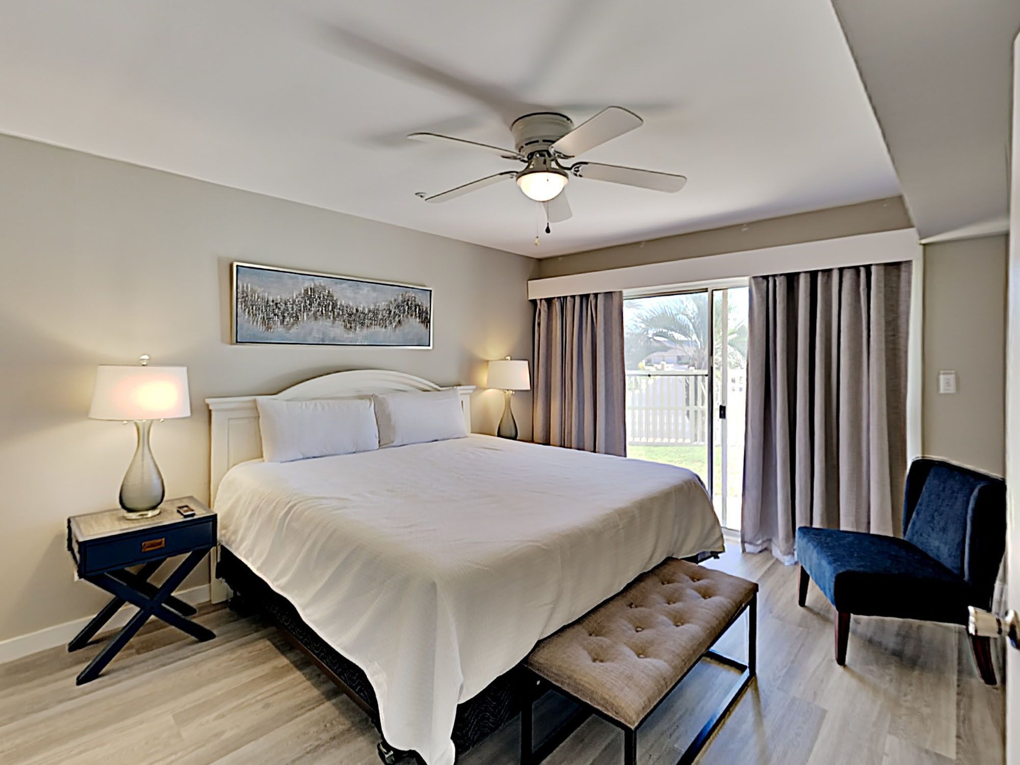 Ein Hotelzimmer mit einem großen Bett, einem Deckenventilator, einer Bank, einem Sessel und Blick ins Freie durch eine Schiebetür.