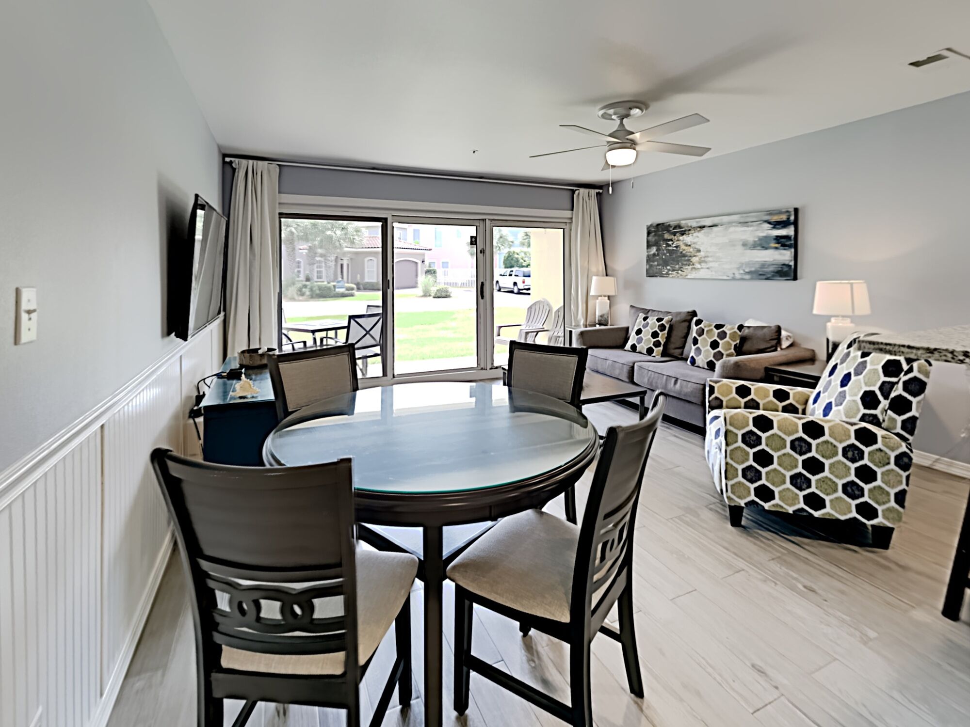 A dining area with a round table and chairs, a sofa with patterned cushions, a ceiling fan, and a view of the outdoors through a glass door.