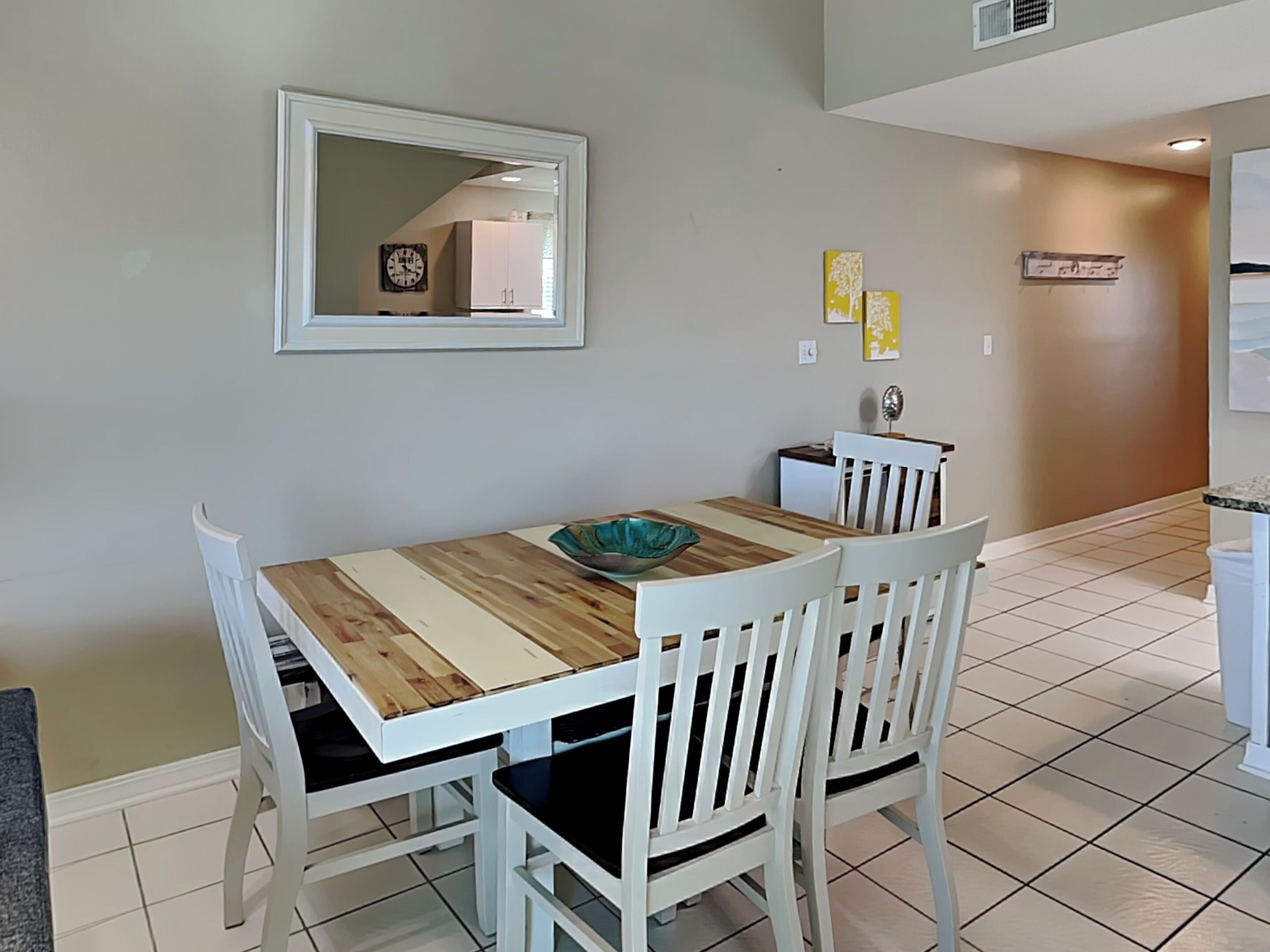 Een eetgedeelte met een houten tafel en witte stoelen, een kleine keuken op de achtergrond en een spiegel aan de muur.