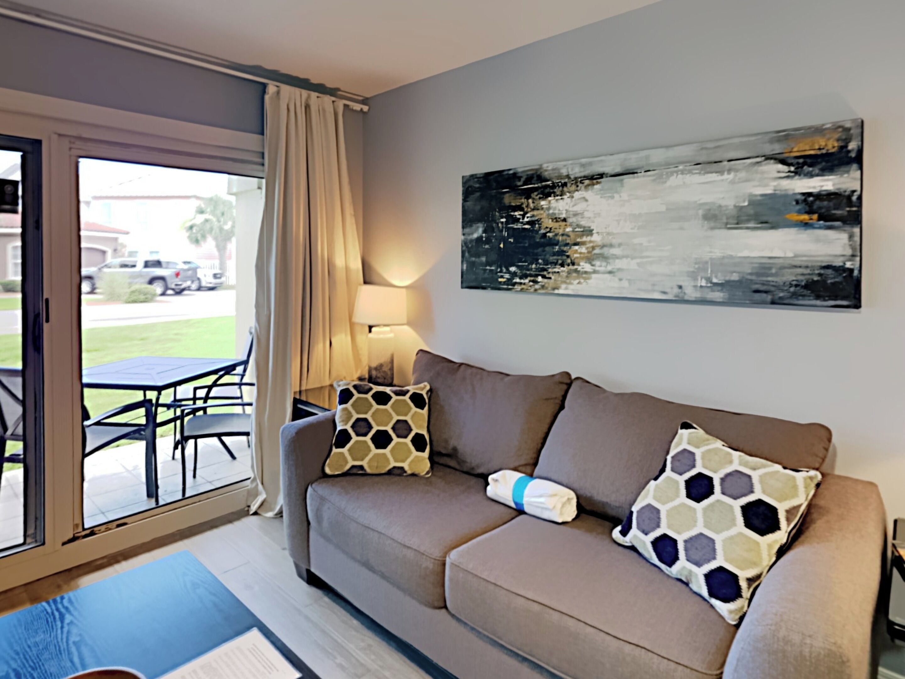Un soggiorno moderno con un divano, due cuscini decorativi a motivi, un grande quadro astratto, un tavolo con sedie e una porta vetrata scorrevole che dà su un’area esterna.