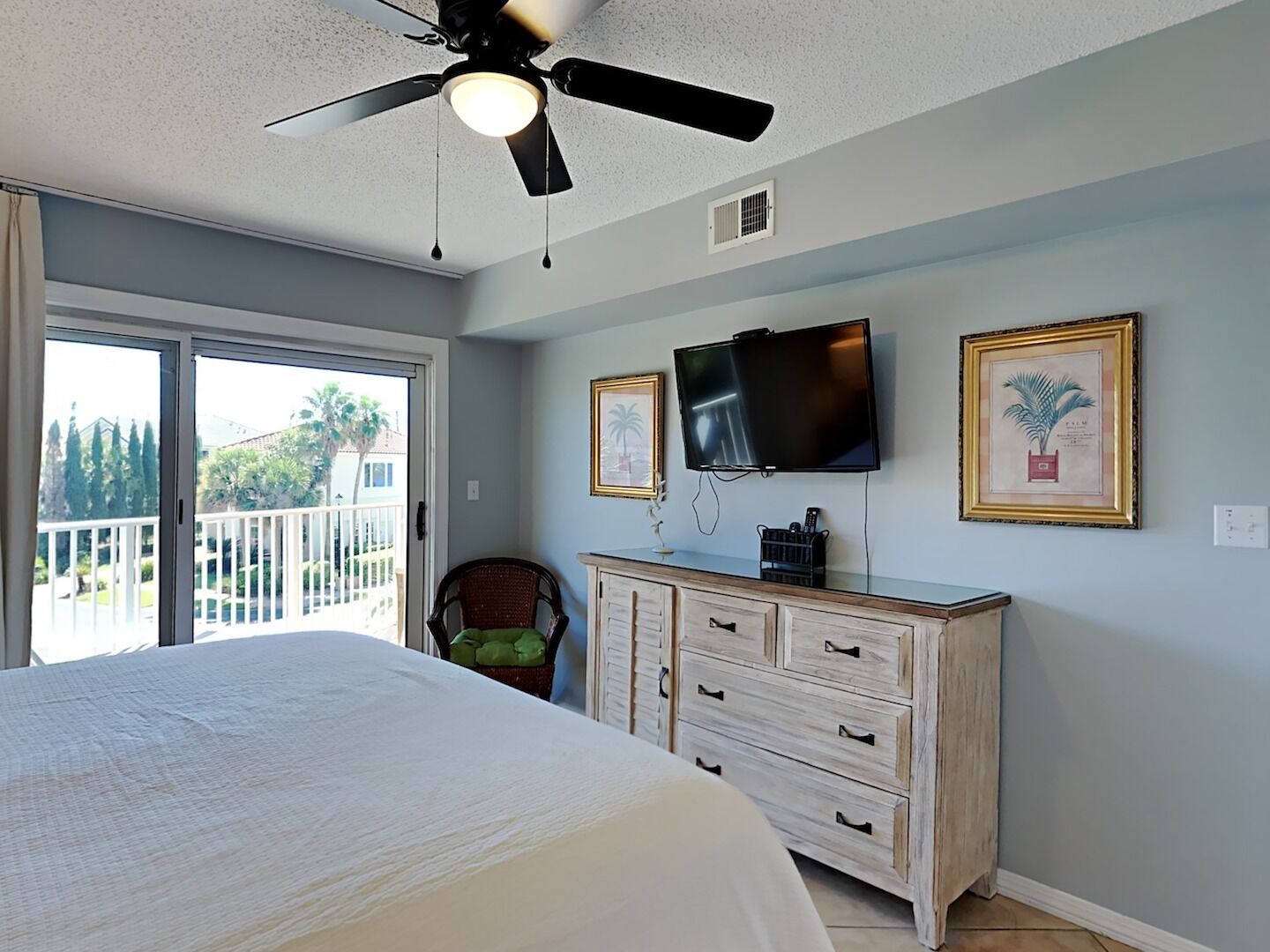 A bedroom with a bed, a ceiling fan, a TV, a dresser, and a balcony with a view of trees and a house.