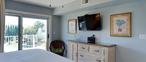 Un dormitorio con cama, ventilador de techo, televisor, cómoda y balcón con vistas a árboles y una casa.
